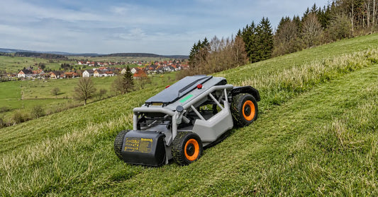 Ein Mowrator-Mähroboter fährt im Frühling einen grasbewachsenen Hang hinauf, während im Hintergrund ein Dorf und sanft geschwungene grüne Felder zu sehen sind.